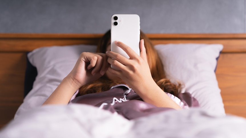 A woman lies in bed, under the covers, messaging on her phone.
