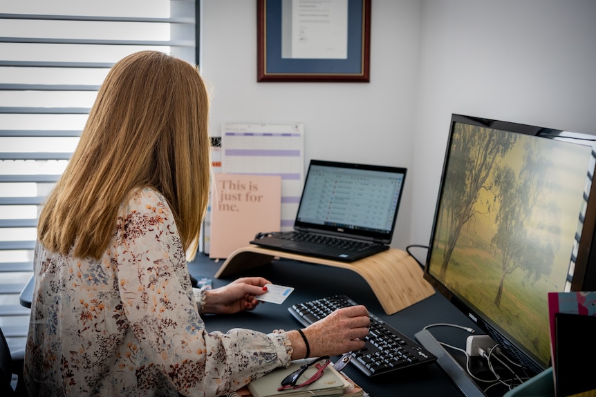 A woman sits at a desk in front of a monitor and laptop