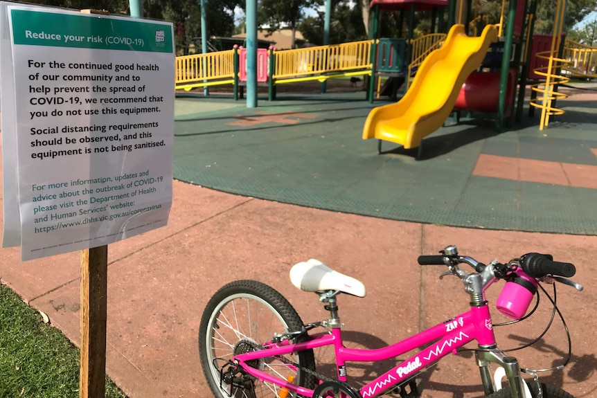 A sign warning about the risks posed by COVID-19 at a children's playground with a yellow slide and pink bicycle.