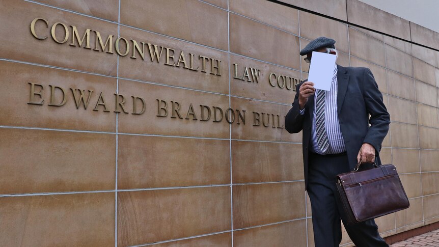 An elderly portly man walks into court wearing a suit and holding a book to hide his face 