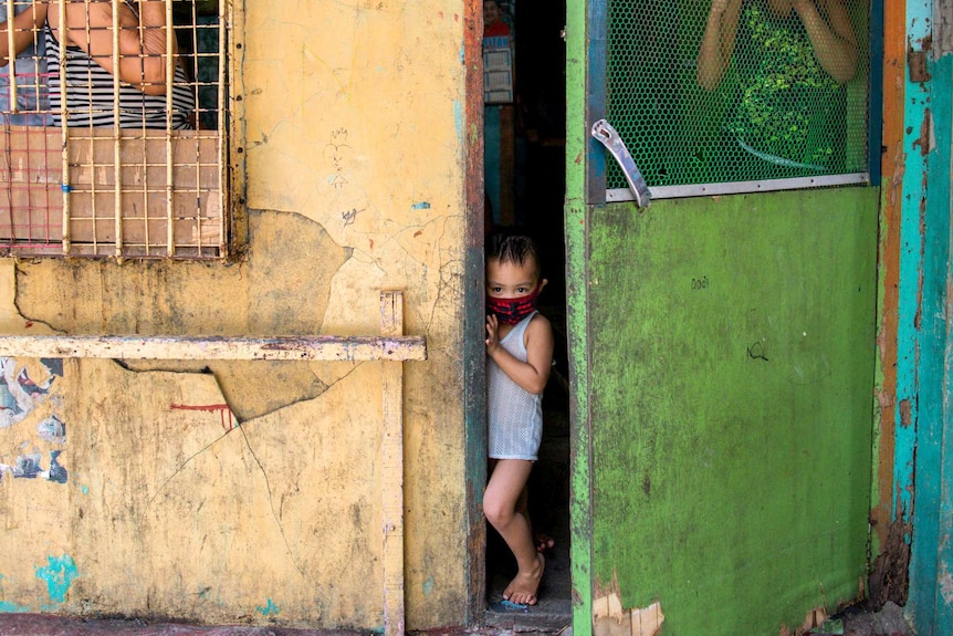 A small child in a face mask looks out a doorway