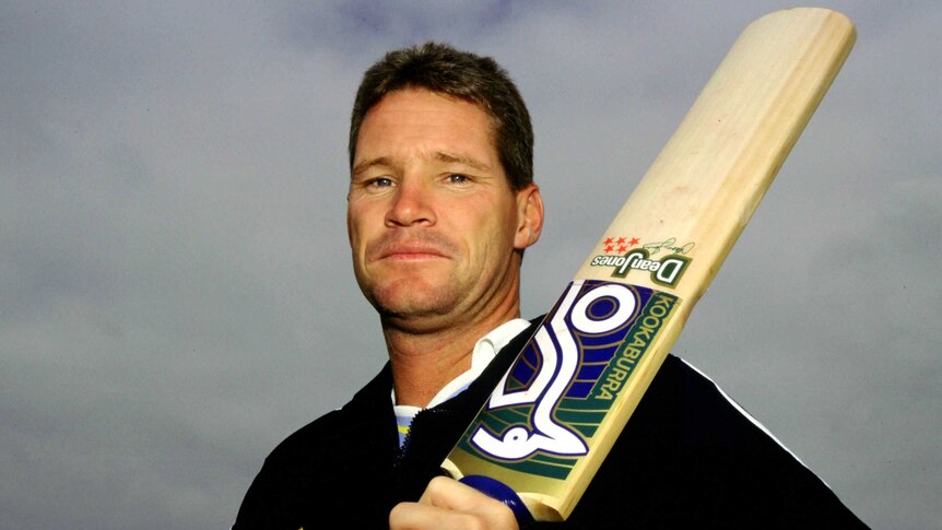An Australian cricketer stands looking down at the camera as he holds a cricket bat over his left shoulder.