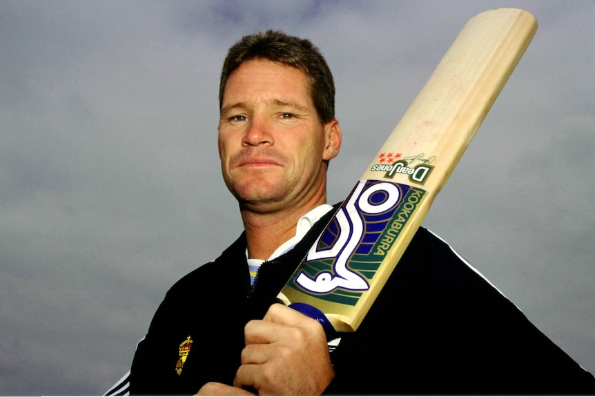 An Australian cricketer stands looking down at the camera as he holds a cricket bat over his left shoulder.