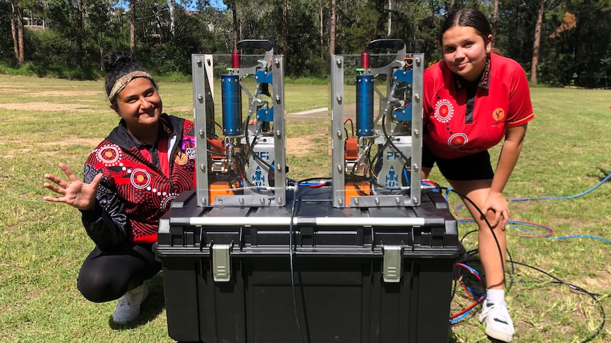 An image of two students with a rocket motor on a school oval with trees in the background