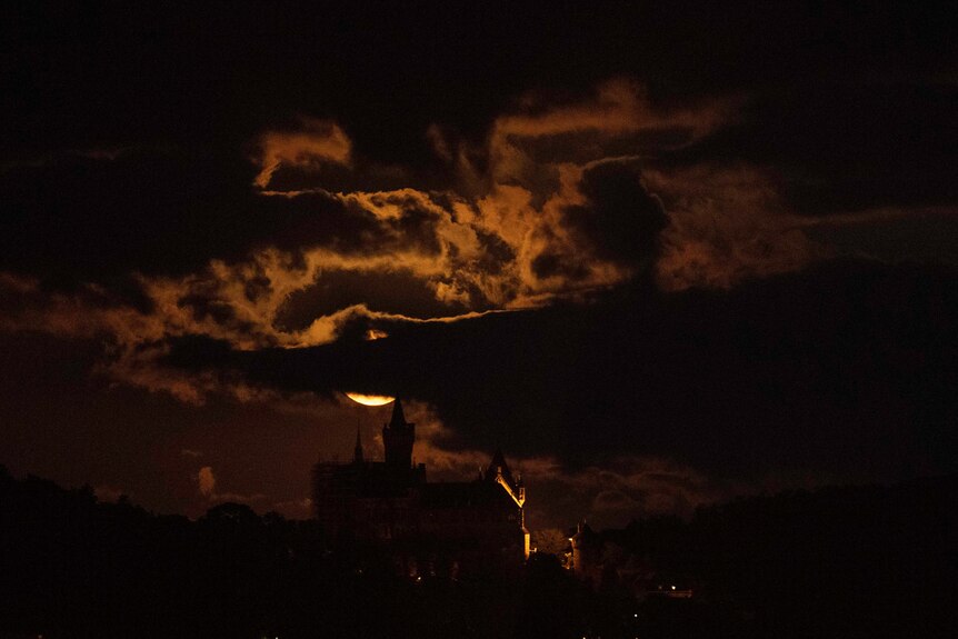 A large moon obscured by clouds rises behind a castle, illuminating clouds and sky in orange light.