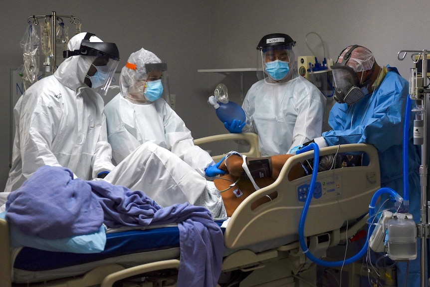 A group of health workers in full PPE surround a man on a hospital bed