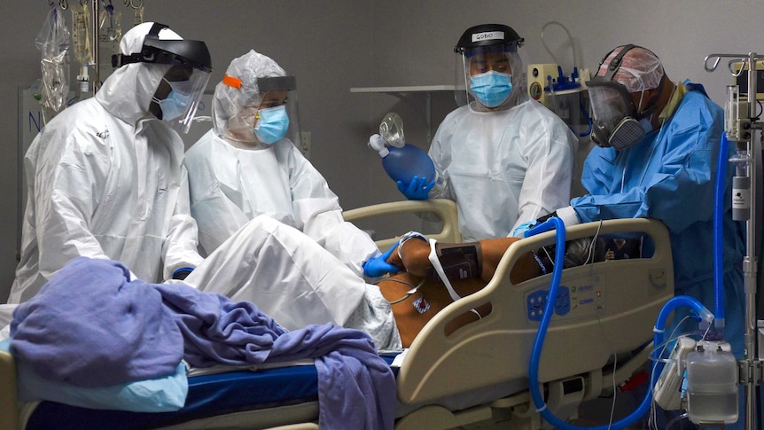A group of health workers in full PPE surround a man on a hospital bed