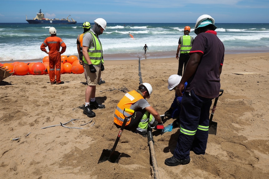 A group of workmen bury a cable underneath sand on a beach. A bit further out to sea, a boat is seen.