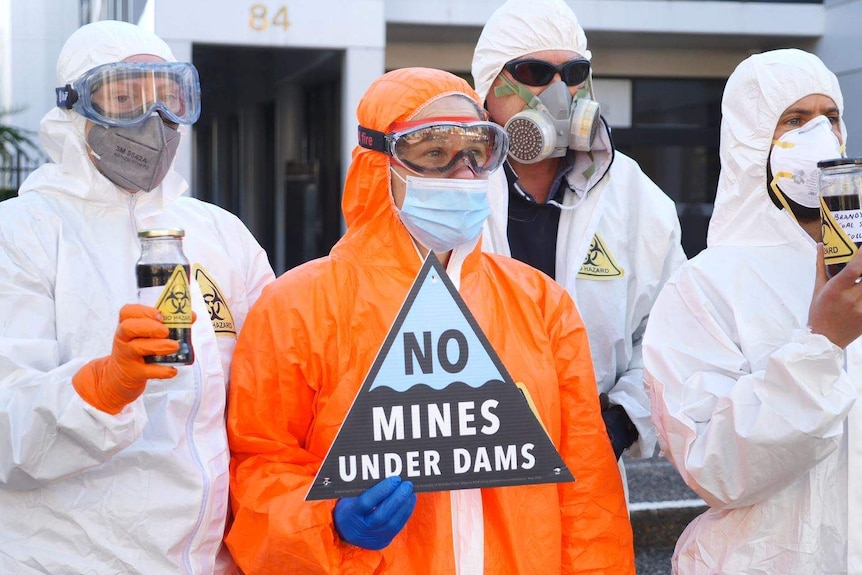 Protesters stand in bio hazard suits, holding glass bottles with sludge in them.