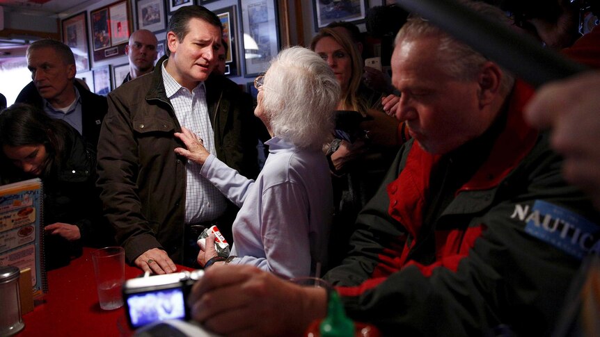 An elderly woman places her hand on the chest of Ted Cruz as they talk while standing at the counter in a diner.