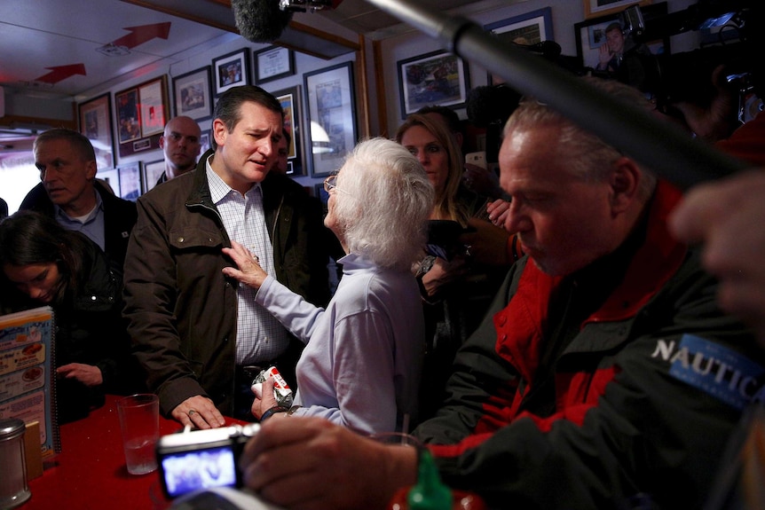 An elderly woman places her hand on the chest of Ted Cruz as they talk while standing at the counter in a diner.