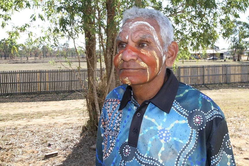 Aboriginal elder with traditional paint on his face stands in front of a tree.