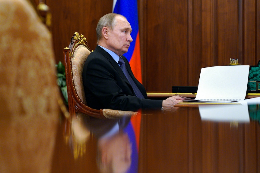 An elderly grey-haired man sits at wood-grain table in a suit with a Russian flag behind.