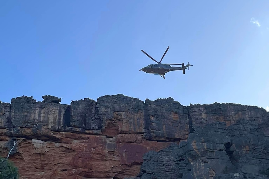 An ambulance helicopter flies over a brown and orange rocky outcrop on a sunny afternoon