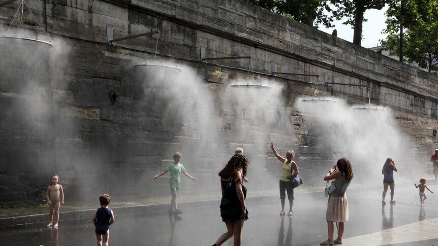 People walk past a cooling water mister system by a stone wall.