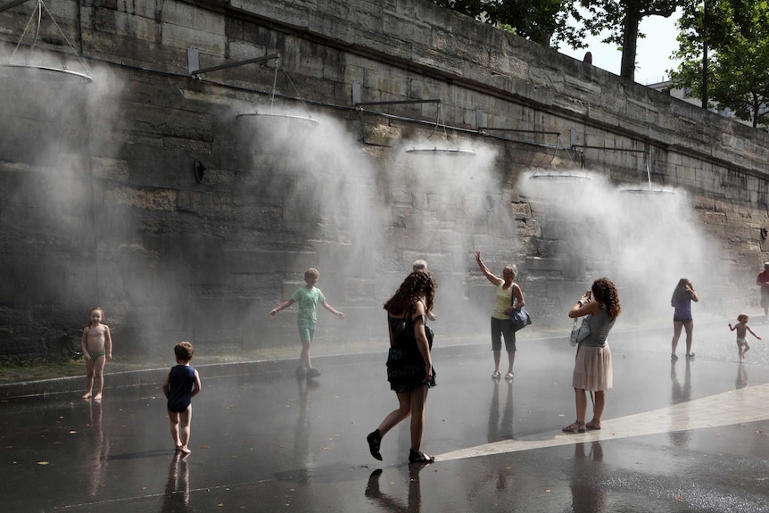 People walk past a cooling water mister system by a stone wall.