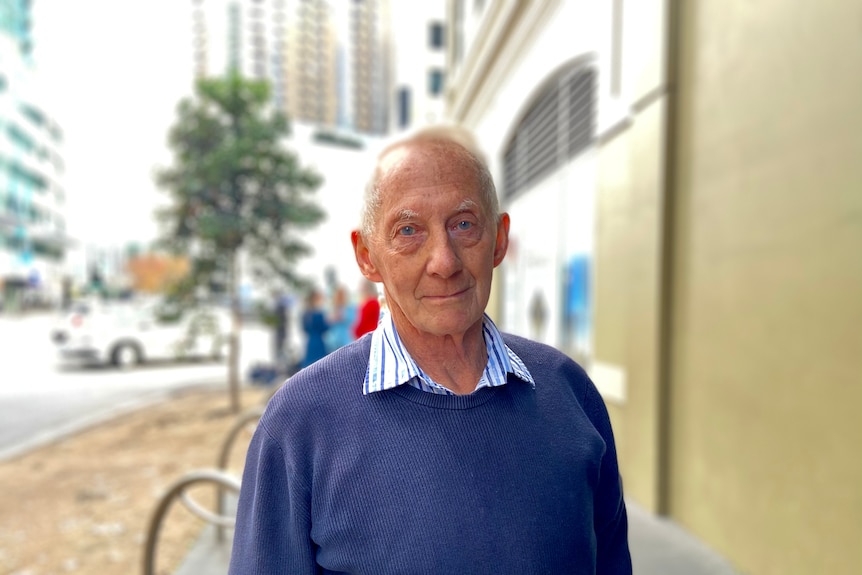 A man in a blue shirt and jumper standing in a street outside.