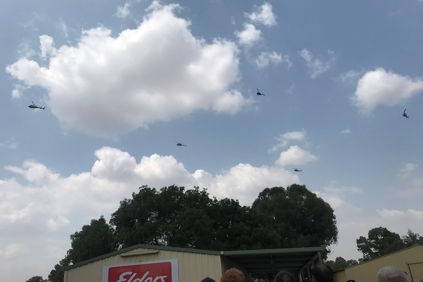 Five helicopters fly over a group of people and buildings.