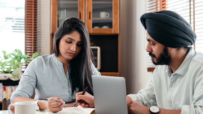 A couple holding pens look at documents while sitting at a kitchen table, in a story about stop putting off making a will.