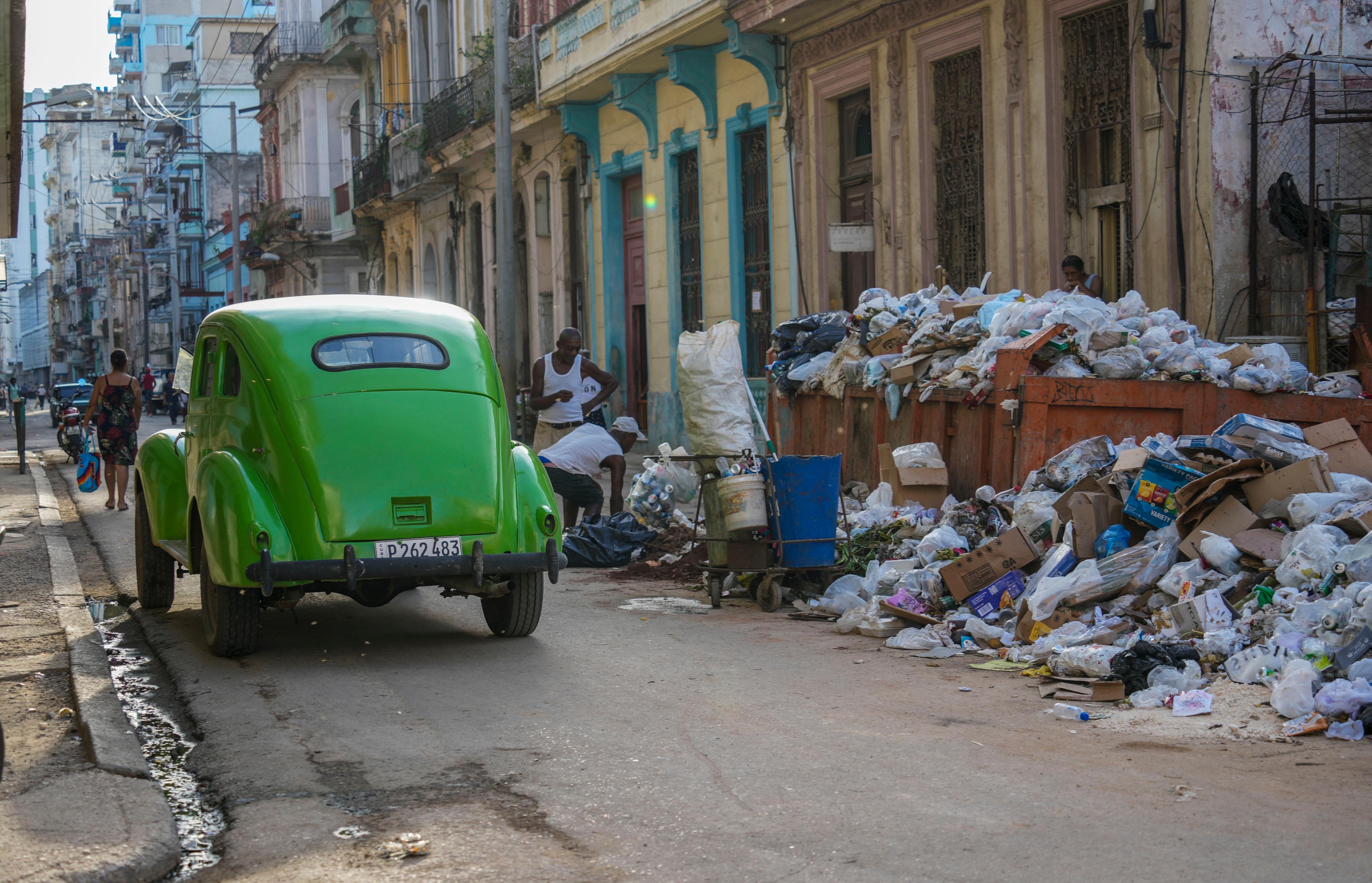A green classic American car driving away from large piles of rubbish flowing out of a container on a road.