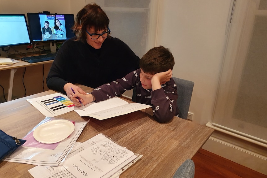 A woman sits next to a young boy who is looking at school books.