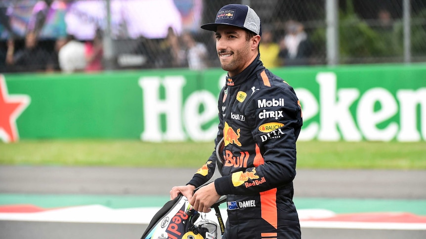 Daniel Ricciardo walks along with a grimace holding his helmet in front of him.