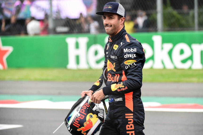 Daniel Ricciardo walks along with a grimace holding his helmet in front of him.