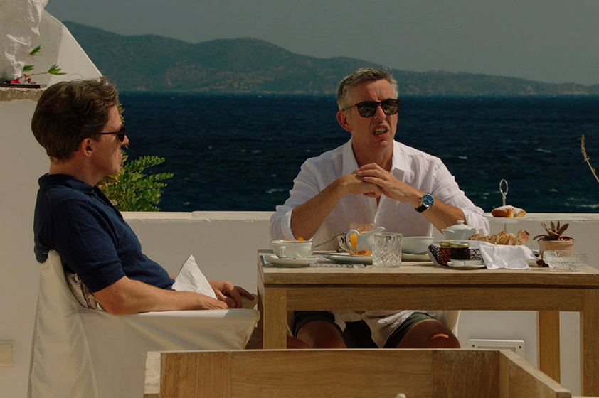 Two middle aged men wear button up shirts, shorts and sunglasses and sit at outdoor dining table by the sea on sunny day.