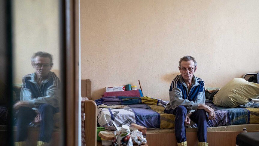 An elderly man sits on a bed in a room.