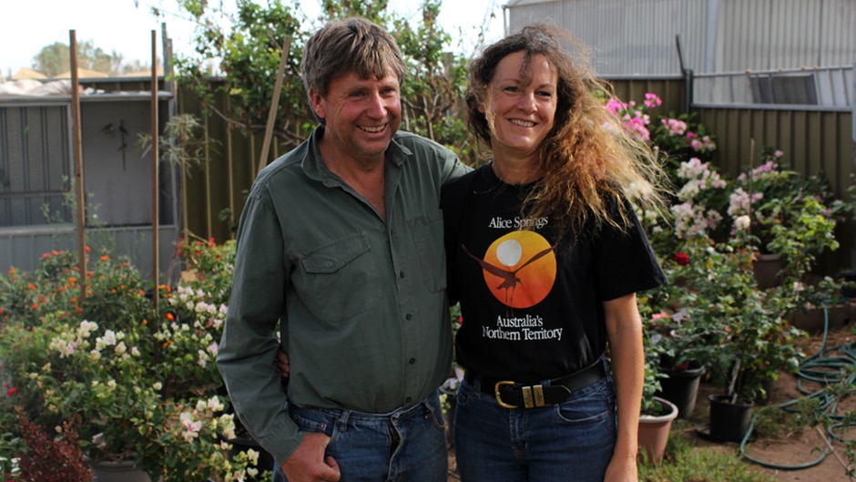 A man in a green shirt and jeans and woman in a black shirt and jeans standing and smiling in front of a garden of flowers.