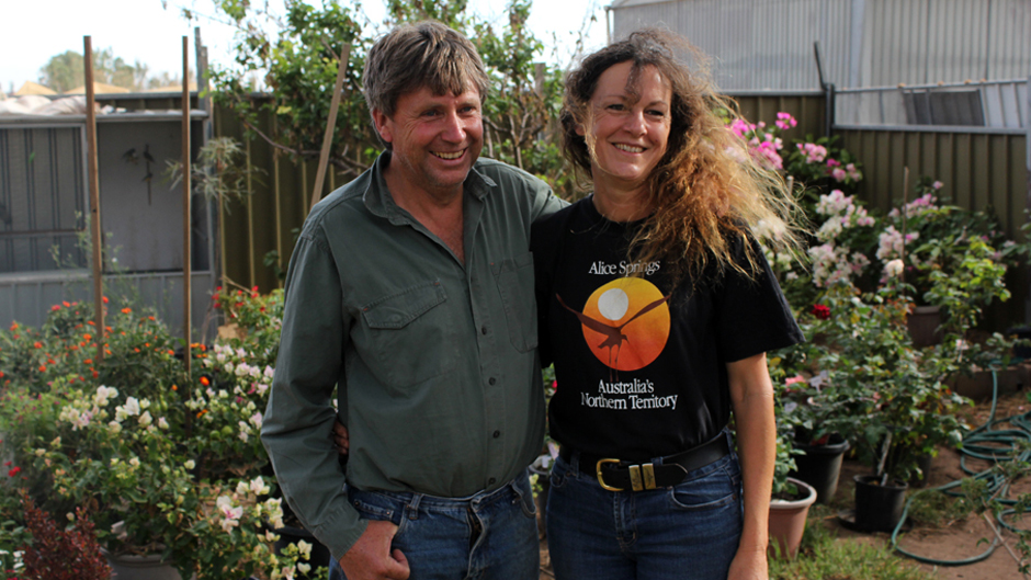 A man in a green shirt and jeans and woman in a black shirt and jeans standing and smiling in front of a garden of flowers.