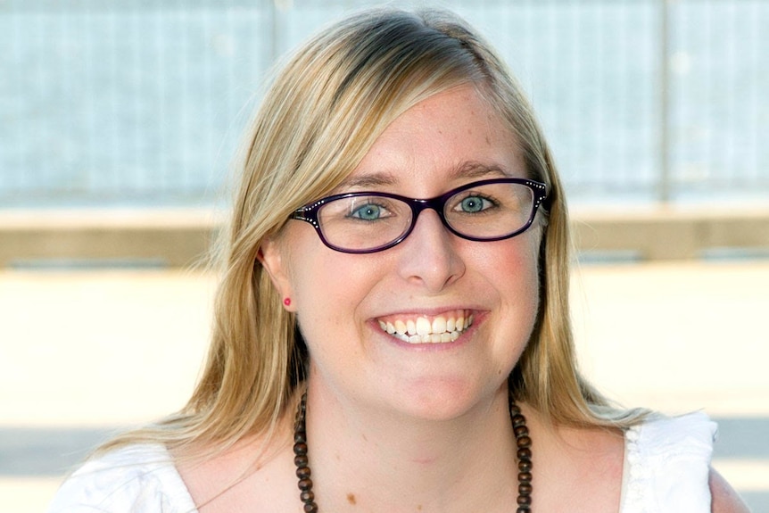 Head shot of widely smiling young woman with blonde hair, blue eyes, in black-rimmed glasses, white top, dark beads around neck.