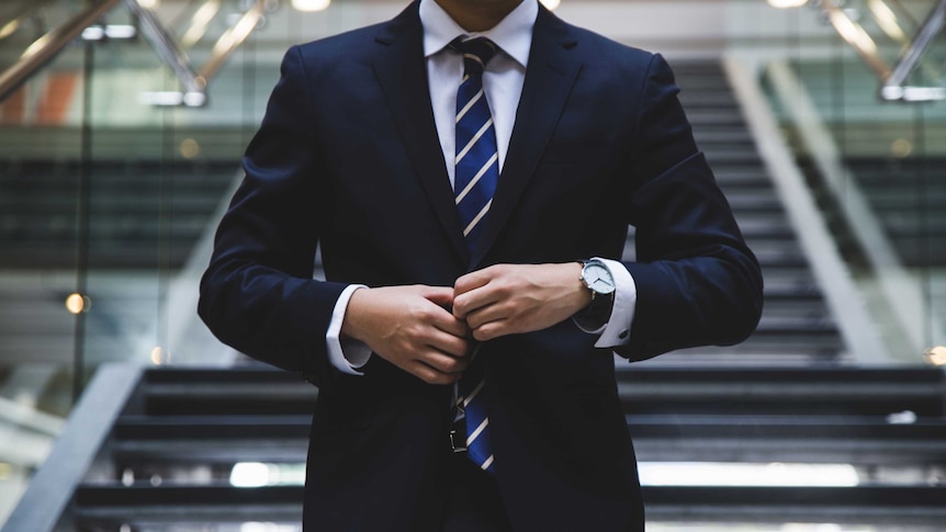 A man does up the top button of a navy suit.