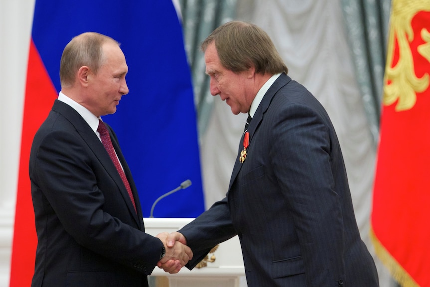 Vladimir Putin shakes hands with a man in a suit, who is bowing slightly before him 