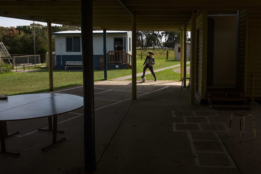 A boy kicks a soccer ball in a patch of light, hopscotch courts in the foreground, several out buildings and bubblers behind.