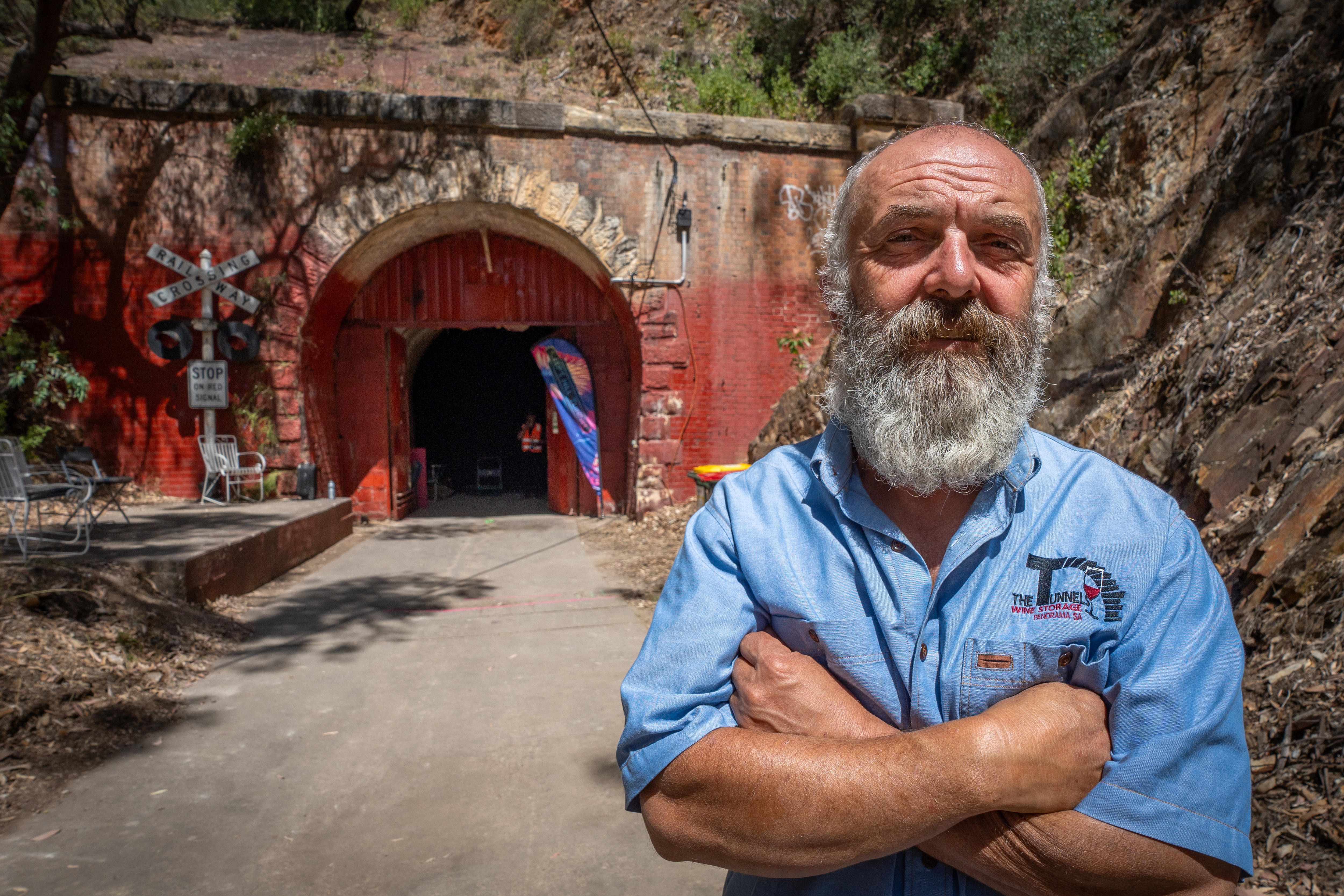 A man with a grey beard,and blue shirt standing with his arms crossed outside a tunnel with a red wall