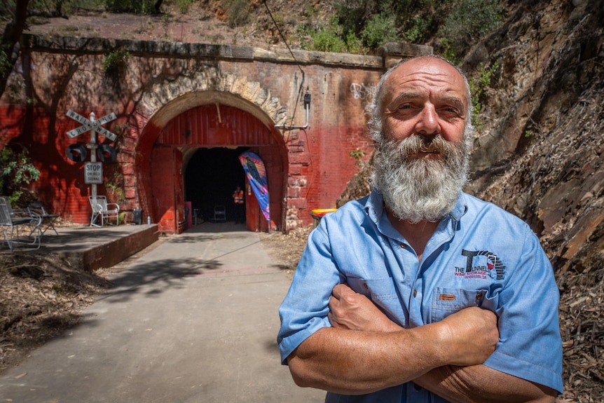 A man with a grey beard,and blue shirt standing with his arms crossed outside a tunnel with a red wall