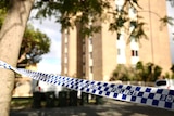 Police tape in the foreground tied to a tree, with a large block of apartments behind.