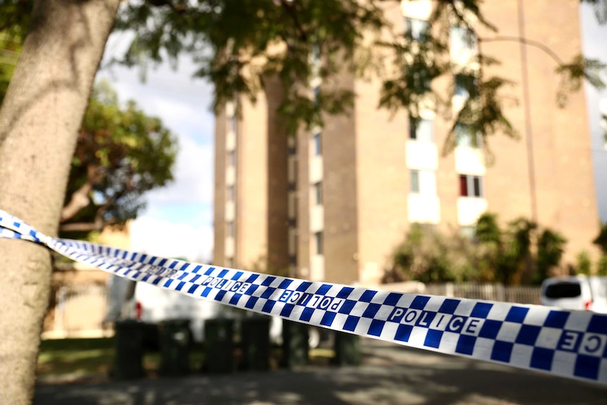 Police tape in the foreground tied to a tree, with a large block of apartments behind.