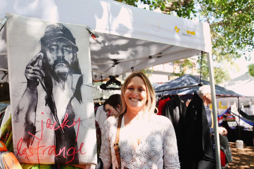 A woman stands in front of a black and white drawing of a man with the words Jacky Le Strange written in red.