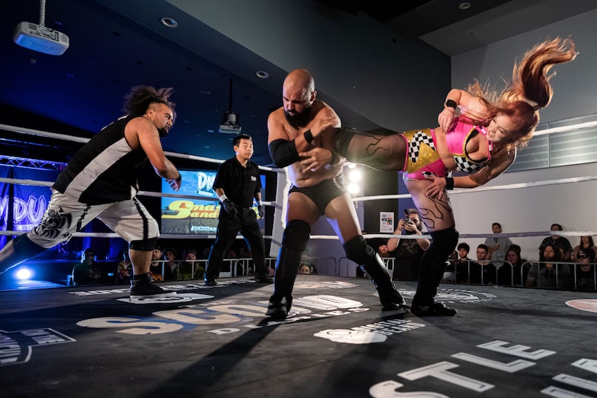 On a boxing ring, a woman in pink lycra fly-kicks a man in black briefs, while another man runs towards them.