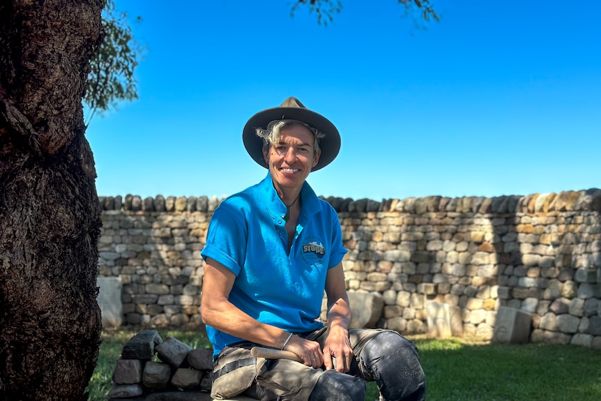 A woman wearing a blue shirt sits in front of a dry stone wall.