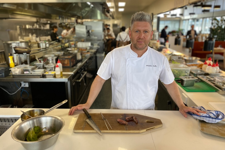 A chef in whites stands in his kitchen with a freshly cut steak.