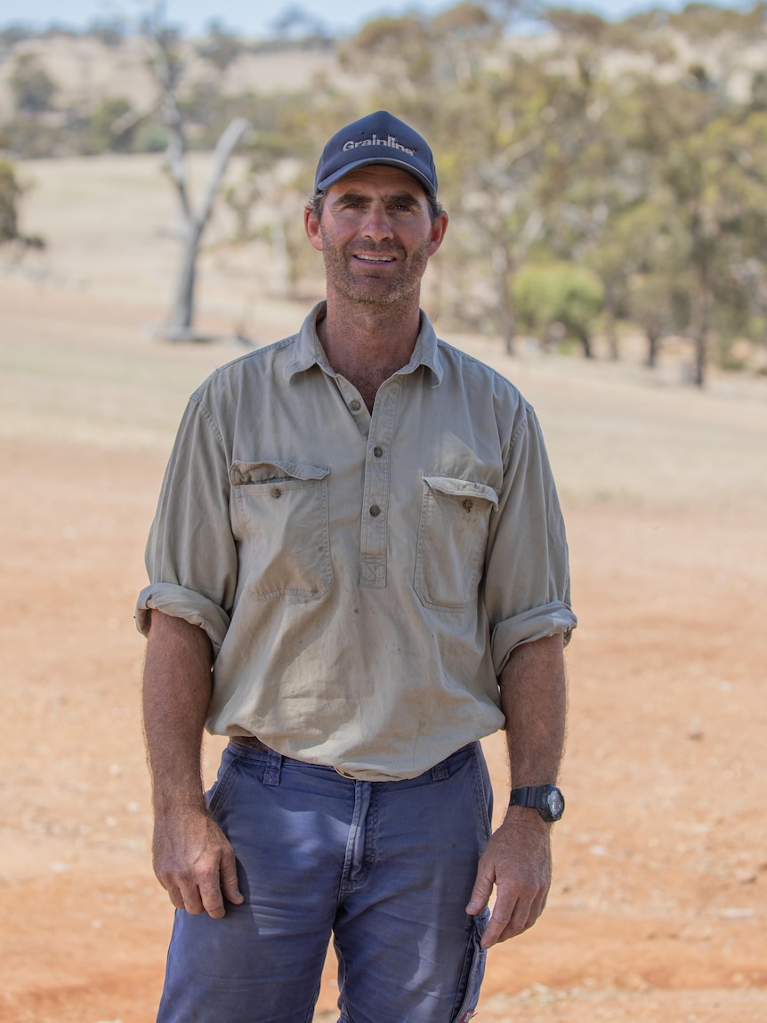 A farmer wearing a baseball cap stands in a red dusty paddock