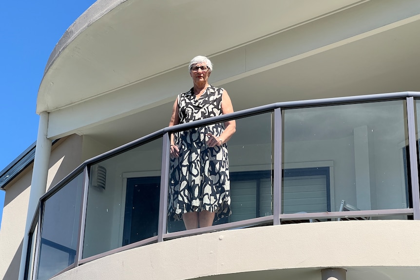 An elderly woman with a black dress and grey hair stands on a balcony.