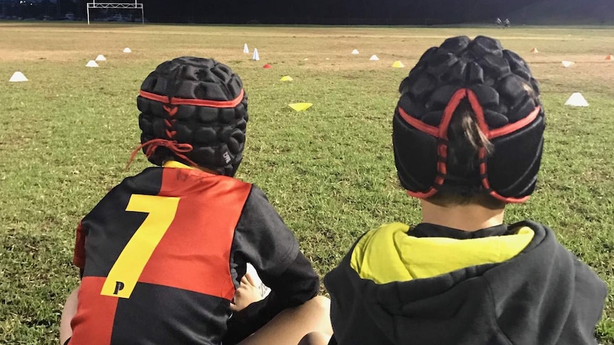 Two children, viewed from the back, sit watching football training.