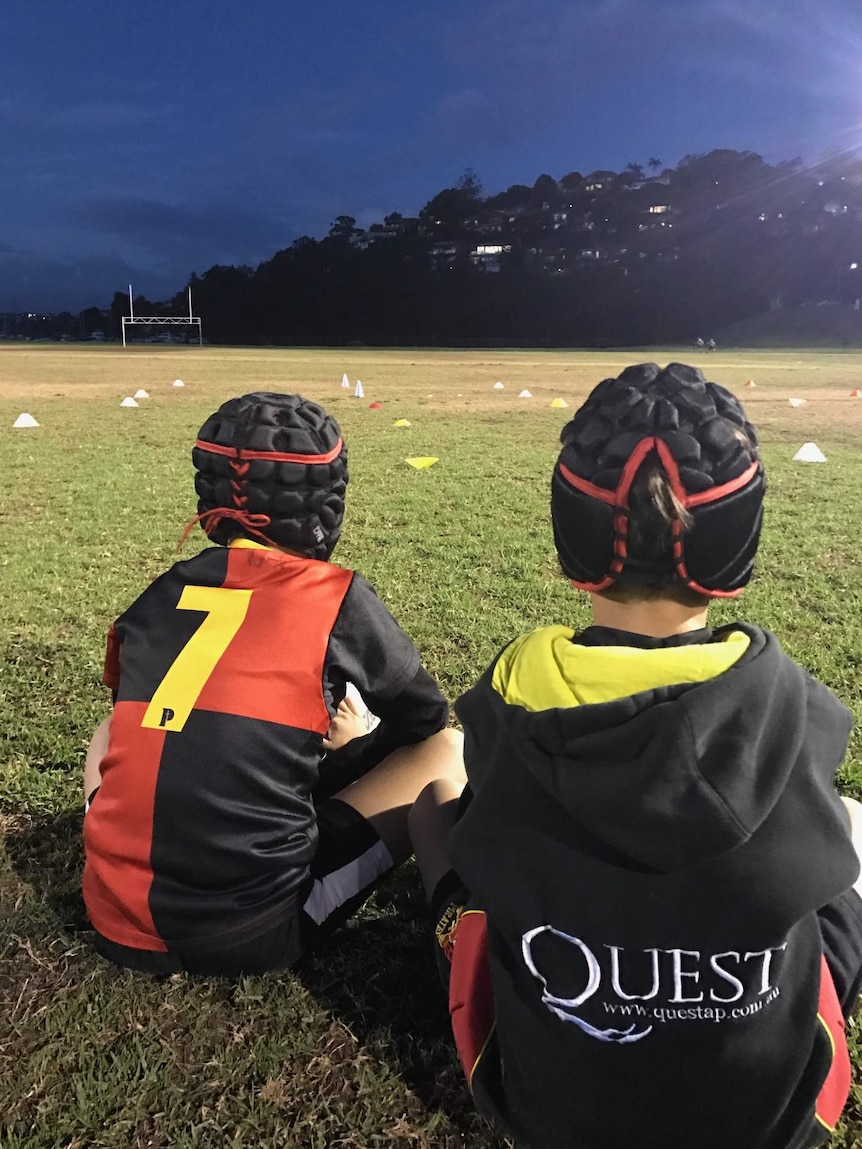 Two children, viewed from the back, sit watching football training.