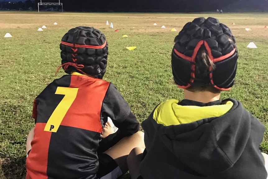 Two children, viewed from the back, sit watching football training.