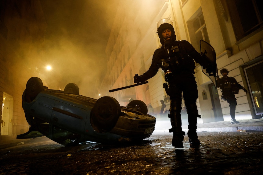 French riot police officers walk next to an overturned vehicle during night under a yellow street light.