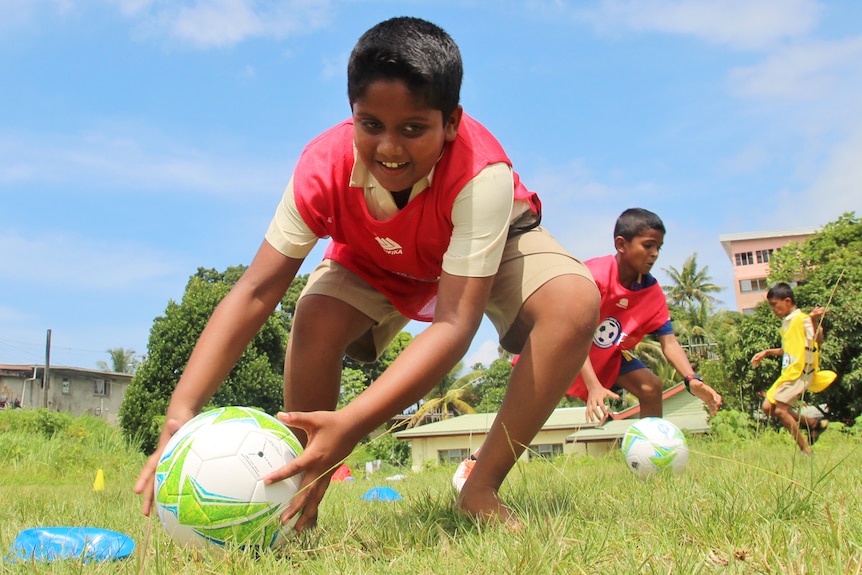 Three boys take part in a race that involves picking up a football and running with it.